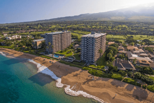Aerial view of beachfront high-rise buildings and homes along a sandy shore with waves, set against a backdrop of green landscape and mountains.