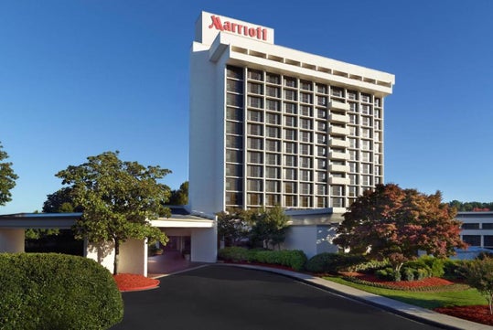Tall white Marriott hotel building with multiple floors and balconies, surrounded by trees and landscaping, under a clear blue sky.