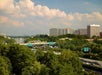 View of a highway surrounded by trees with multiple green road signs, leading toward a cluster of mid-rise office buildings under a partly cloudy sky.