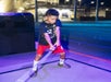 A young boy stands on a lit path holding a glowing toy lightsaber, wearing a black shirt, red shorts, and mismatched socks. An illuminated building is visible in the background.