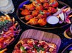 A table with spicy chicken wings, a platter of fries with vegetables and herbs, a wooden board with sliced pizza, and a cold drink.