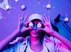 Person lying on the floor, puckering lips, holding two golf balls over eyes. Wearing a cap and surrounded by golf balls and a club. Lit with pink and blue lights.