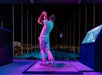 Person in a white outfit swinging a golf club at an illuminated driving range at night, surrounded by screens and targets.