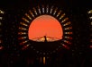 A silhouetted performer stands on stage in front of a large circular orange and yellow light display with triangular patterns, resembling a sunset.