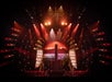 A theatrical stage is lit with red and orange lights, featuring acrobats performing on aerial silks, geometric set pieces, and a vivid sunset backdrop.