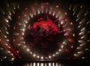 Two acrobats perform on a large rotating metal apparatus shaped like an infinity symbol, with red and orange lights and geometric patterns in the background.