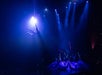 Aerial performer suspended from a hoop under blue and purple stage lights, with dancers in long flowing dresses posing on stage below.