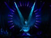 A performer balances on one leg atop a transparent bowl on stage, surrounded by blue spotlights and geometric light patterns, with an audience and other performers visible.