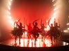 Seven dancers in traditional costumes perform a choreographed routine with sticks on a stage, silhouetted against dramatic red lighting.