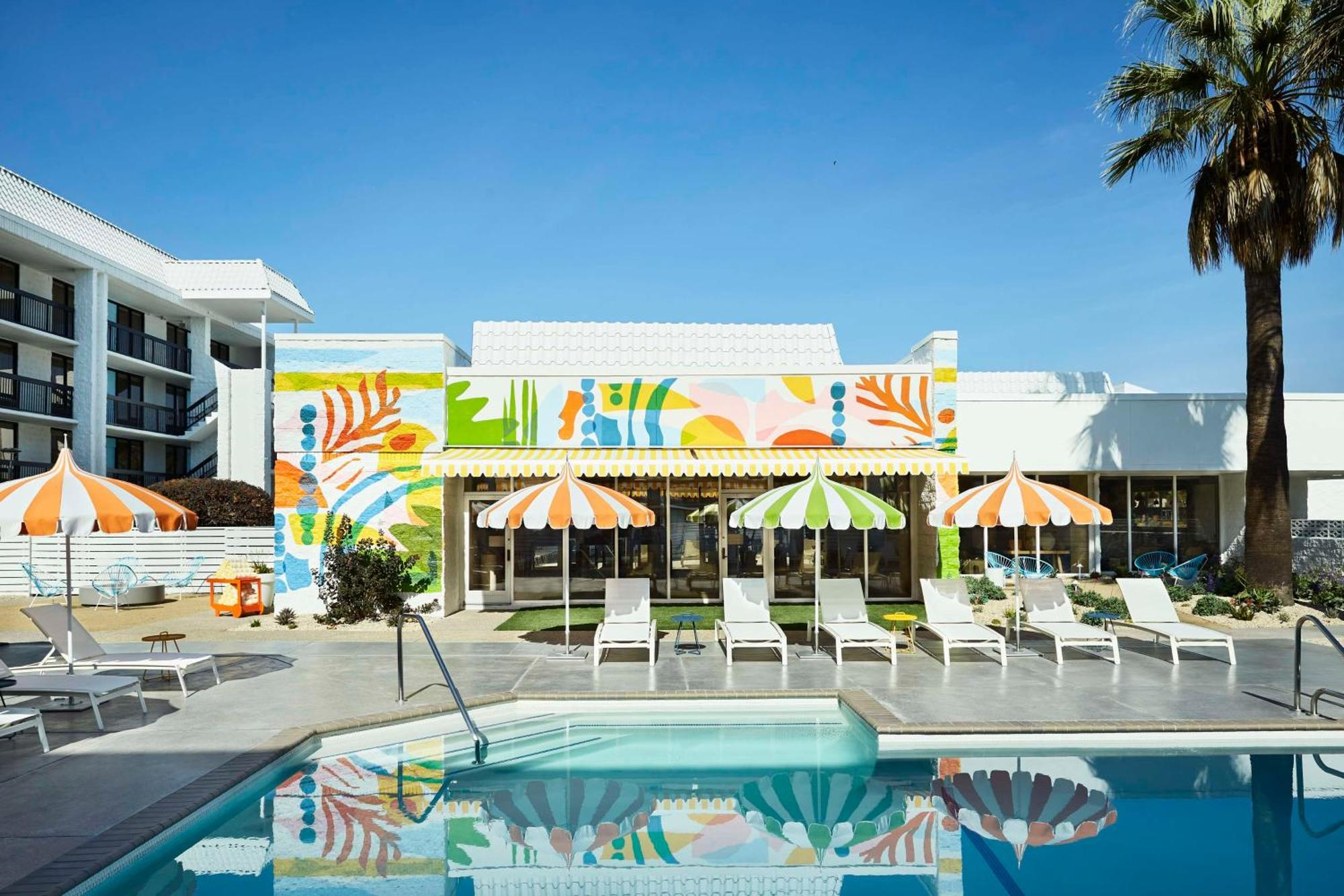 Outdoor pool area with white lounge chairs, striped orange umbrellas, and a colorful mural on the building under a clear blue sky.