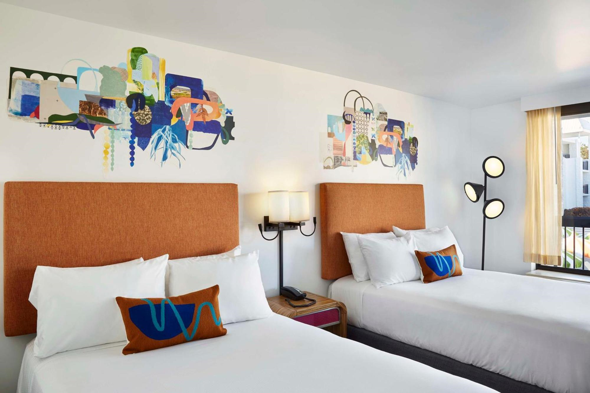 Hotel room with two double beds, orange headboards, abstract wall art above each bed, white bedding, decorative pillows, a wall lamp, and a modern floor lamp by the window.