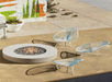 Three blue and white modern chairs are arranged around a round concrete fire pit on a sandy patio, with a colorful mural and plants in the background.