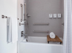 Accessible bathroom with a bathtub, multiple grab bars, a handheld showerhead, a wooden bath bench, a rolled white towel, and wall-mounted soap dispensers.