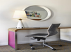 A modern office chair sits at a wooden desk with a white lamp and a round mirror on the wall above, reflecting part of a colorful artwork.