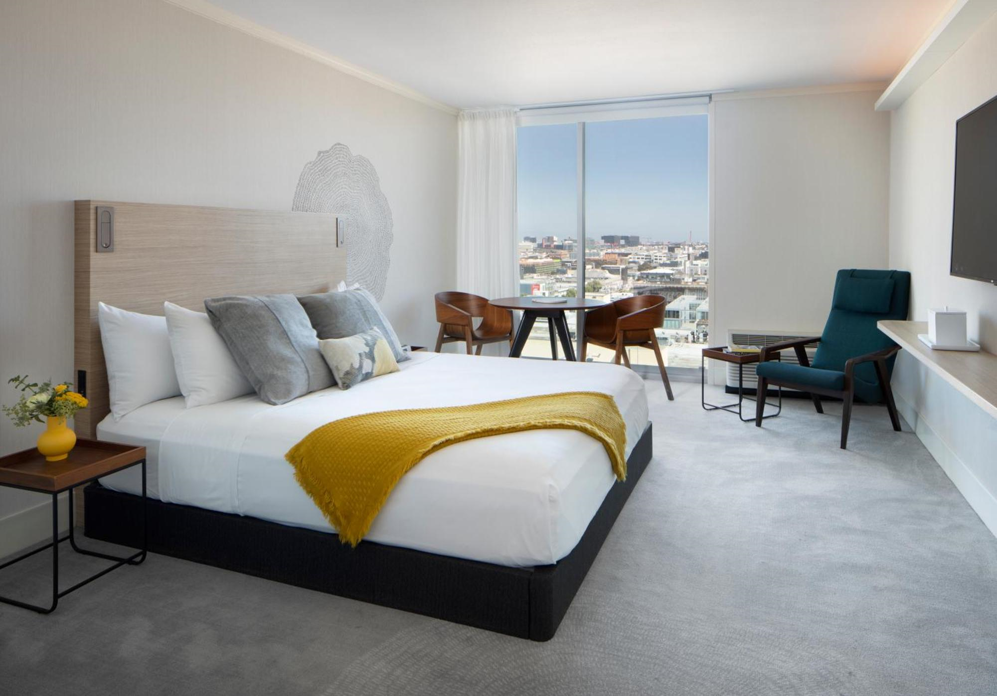 Modern hotel room with a king bed, yellow throw blanket, lounge chair, table with two chairs, large window with city view, and neutral decor.