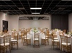 A banquet hall with round tables covered in white tablecloths, set with floral centerpieces and wooden chairs, ready for an event.