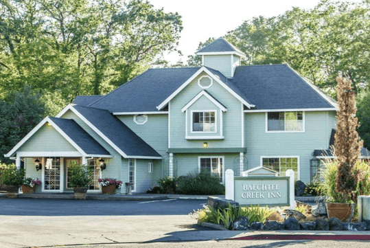 The exterior of the Baechtel Creek Inn with light green siding and grey roof and lots of landscaping on a sunny day.