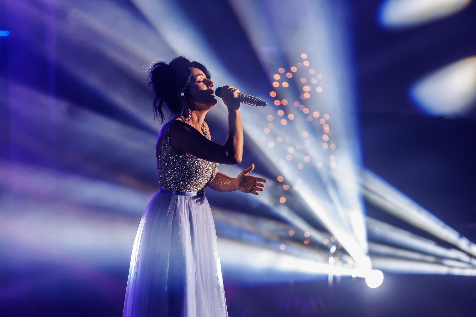 A woman in a sparkling dress sings into a microphone on stage, with dramatic lighting and beams of light in the background.