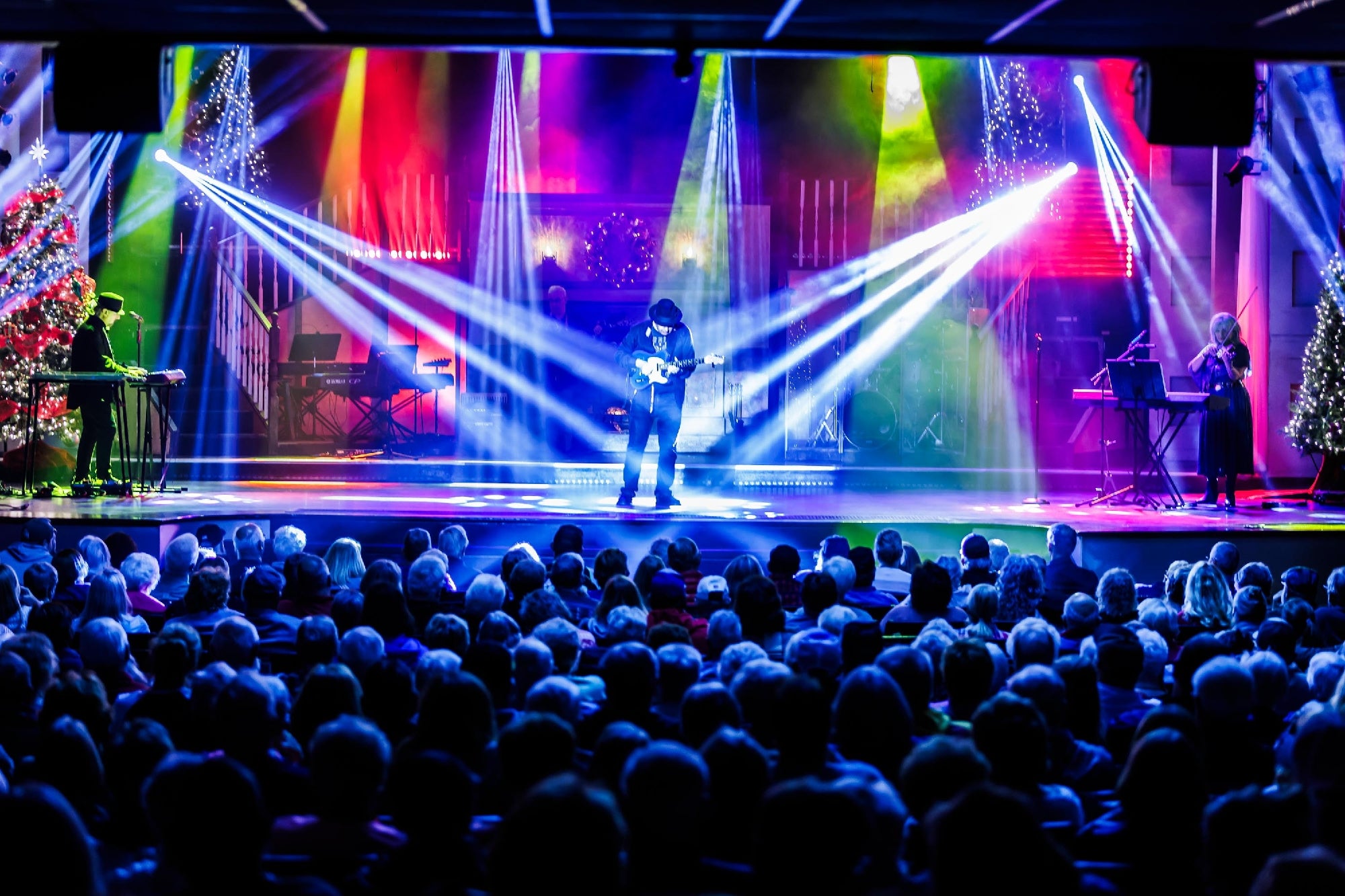 A musician performs on stage with colorful lights and fog, backed by a band, in front of a seated audience in a theater.