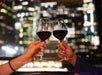Two guests sheeting their glasses of red wine together with the city lights in the background on the Signature Spirit of Baltimore Dinner Cruise.