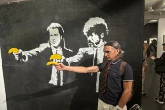 A man poses in front of a mural depicting two figures holding bananas instead of guns, mimicking the gesture while holding a real banana in his hand.