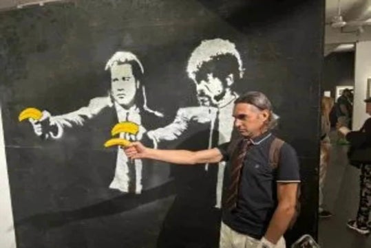 A man poses in front of wall art depicting two figures holding bananas instead of guns, mimicking their gesture with a real banana in his hand.