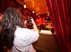 Person with long hair takes a photo of an empty, ornate dining room with red curtains, round tables, and a television on the far wall.