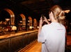 A woman with hair in a low bun takes a photo of a bar with arched mirrors and shelves of bottles, wearing a white shirt that says "TOGETHER" on the back.