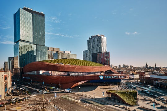 Barclays Center arena with a green roof, surrounded by modern high-rise buildings and city streets under a clear blue sky.