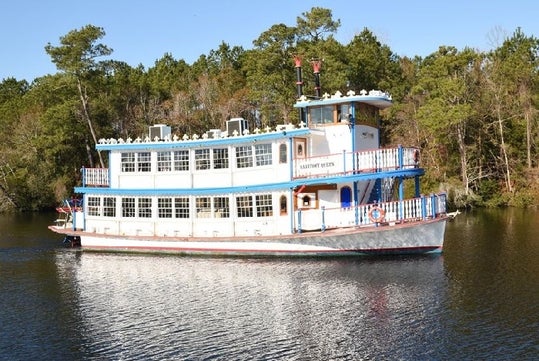 A white, red and blue double-decker river boat with the "Barefoot Queen" in the water with a line a trees behind it on a sunny day.
