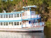 The front of the Barefoot Queen Riverboat Cruise in the water with the sun shining on it and trees in the background.