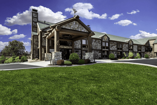 The front exterior and covered driveway of the Bass Pro Shops Angler’s Lodge with green grass in front of it and a blue cloudy sky overhead.