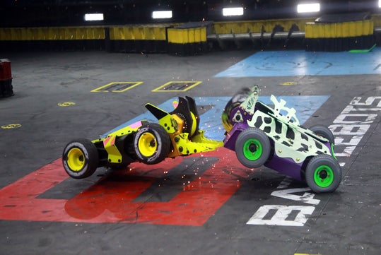Two combat robots collide mid-air in an arena, with sparks flying. They have colorful designs on a marked floor labeled "BATTLEBOTS.