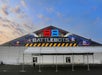 Front view of a large tent with "BattleBots" signage, displaying robotic combat imagery. The structure has a white exterior with a black and yellow striped design.