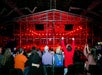 People seated in an arena watch a robot competition inside a red-lit cage, with one person raising their arms in excitement.