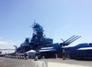 A large battleship is docked at a pier under a clear sky, with visitors walking nearby and several tents and signs visible along the waterfront.