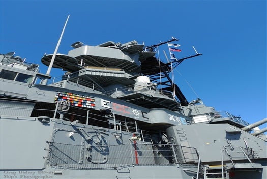 Gray military ship superstructure with communication equipment, flags, and symbols displayed on the side, set against a clear blue sky.