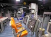 A control room with several control panels, switches, and chairs in orange and blue. The room has various technical equipment and screens, with a desk and several chairs along the side.