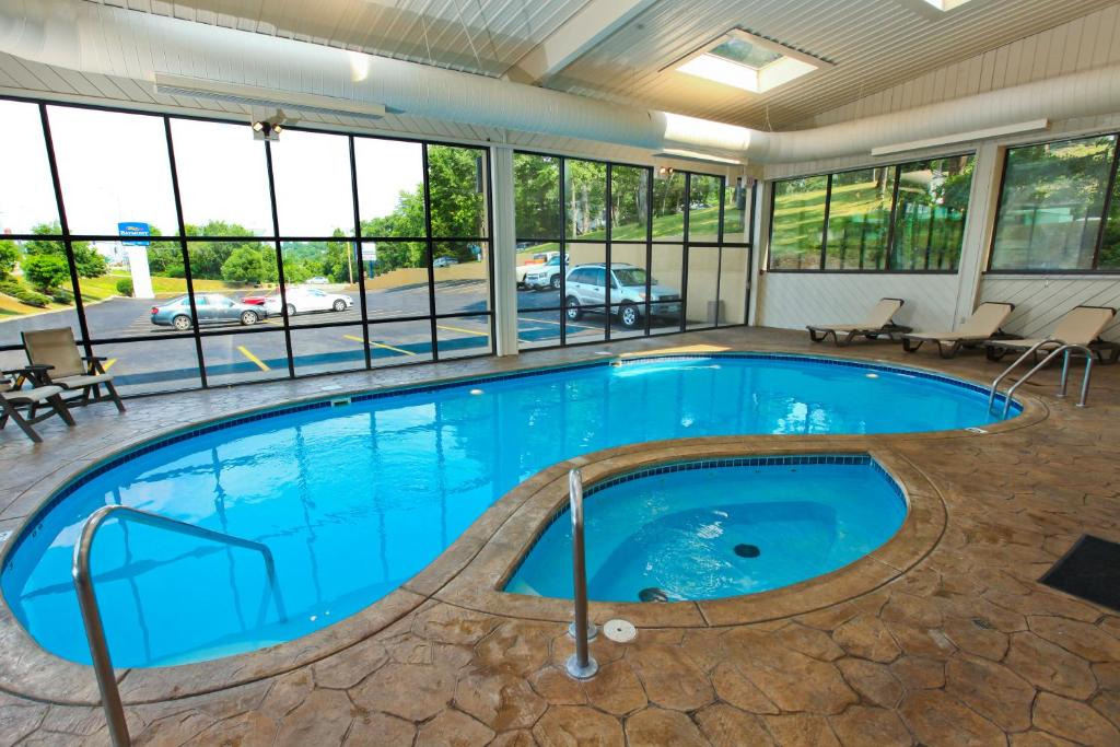 Indoor swimming pool with clear water and stone-patterned deck surrounded by lounge chairs. Large windows provide a view of trees and a parking lot with cars.
