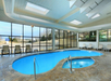 Indoor pool area with a small main pool and a hot tub, surrounded by lounge chairs. Large windows offer a view of the outside.