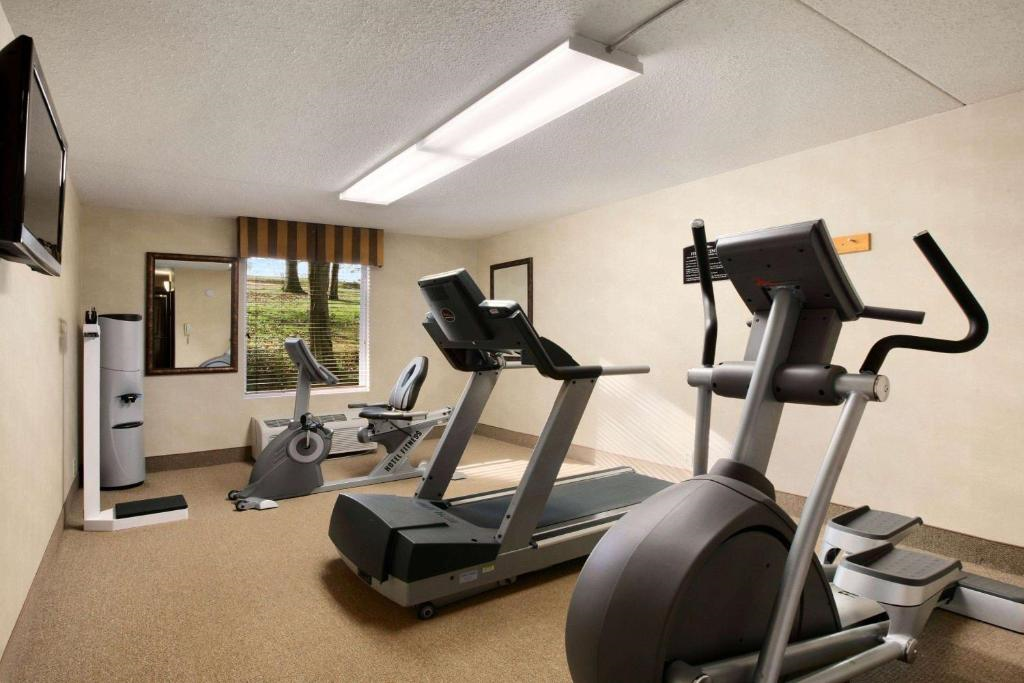 Small gym room with treadmills, an exercise bike, and a standing fan. There is a wall-mounted TV and a window with blinds. The room has beige walls and carpeted floor.