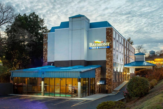 Exterior view of Baymont Inn & Suites, a multi-story hotel with a blue roof, surrounded by trees and a parking area.