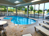 Indoor swimming pool surrounded by lounge chairs, tables, and large windows with outdoor view.