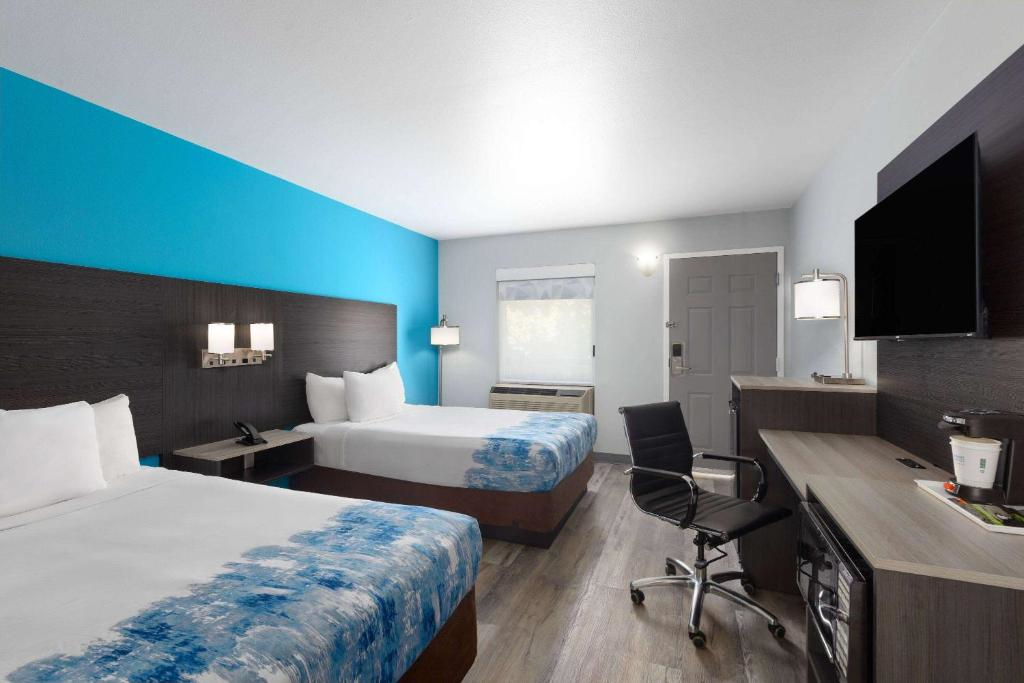 Hotel room with two double beds, a desk with a chair, wall-mounted TV, and bright blue accent wall. Room features modern decor and a window with a view.
