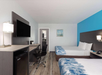 Hotel room with two double beds, a desk with a chair, wall-mounted TV, mini-fridge, and a lamp. Walls are white and blue, and there's a door leading to a bathroom.