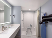 Accessible bathroom with a walk-in shower featuring a shower chair, a white countertop with a sink, toiletries, an iron on a shelf, and some hangers on a rack.