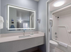 Hotel bathroom with a backlit mirror, sink, toiletries, and a hairdryer. Adjacent to the vanity, there's a white bathtub and shower with a curtain rod.