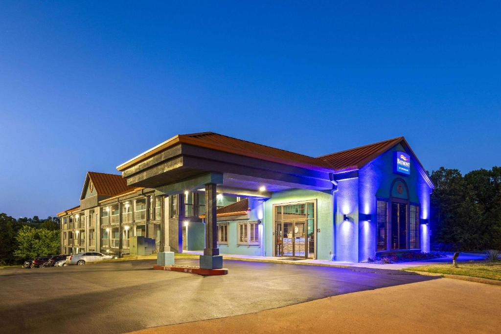 Hotel entrance at dusk with bright blue accent lighting, featuring a covered driveway and multiple glass doors. The building exterior is modern, surrounded by a paved parking area and greenery.