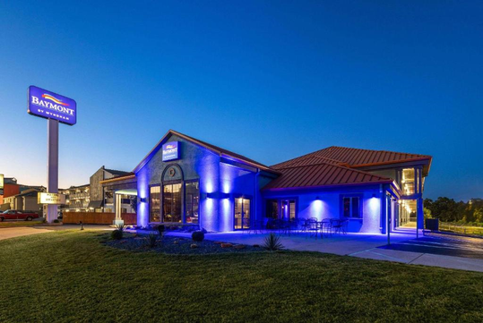 Hotel exterior illuminated with blue lights during twilight, featuring a Baymont by Wyndham sign. Lawn and outdoor seating are visible in the foreground.