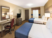 Hotel room with two double beds, a desk with a chair, a TV on a dresser, and a window with curtains. The room has beige walls and carpet flooring.